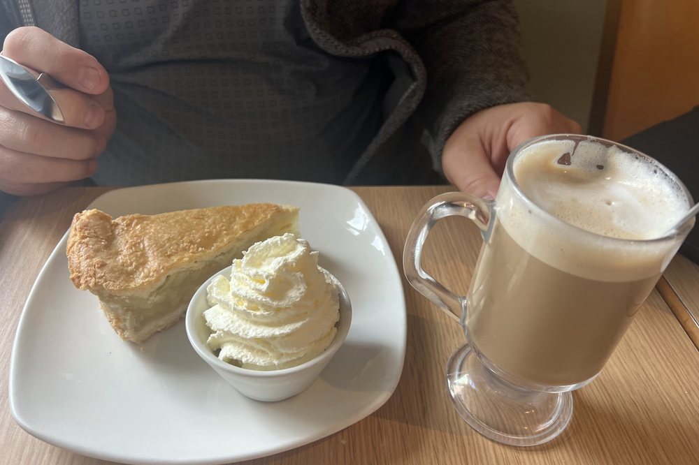 A slice of apple tart with whipped cream beside a tall glass latte at Cates Café.