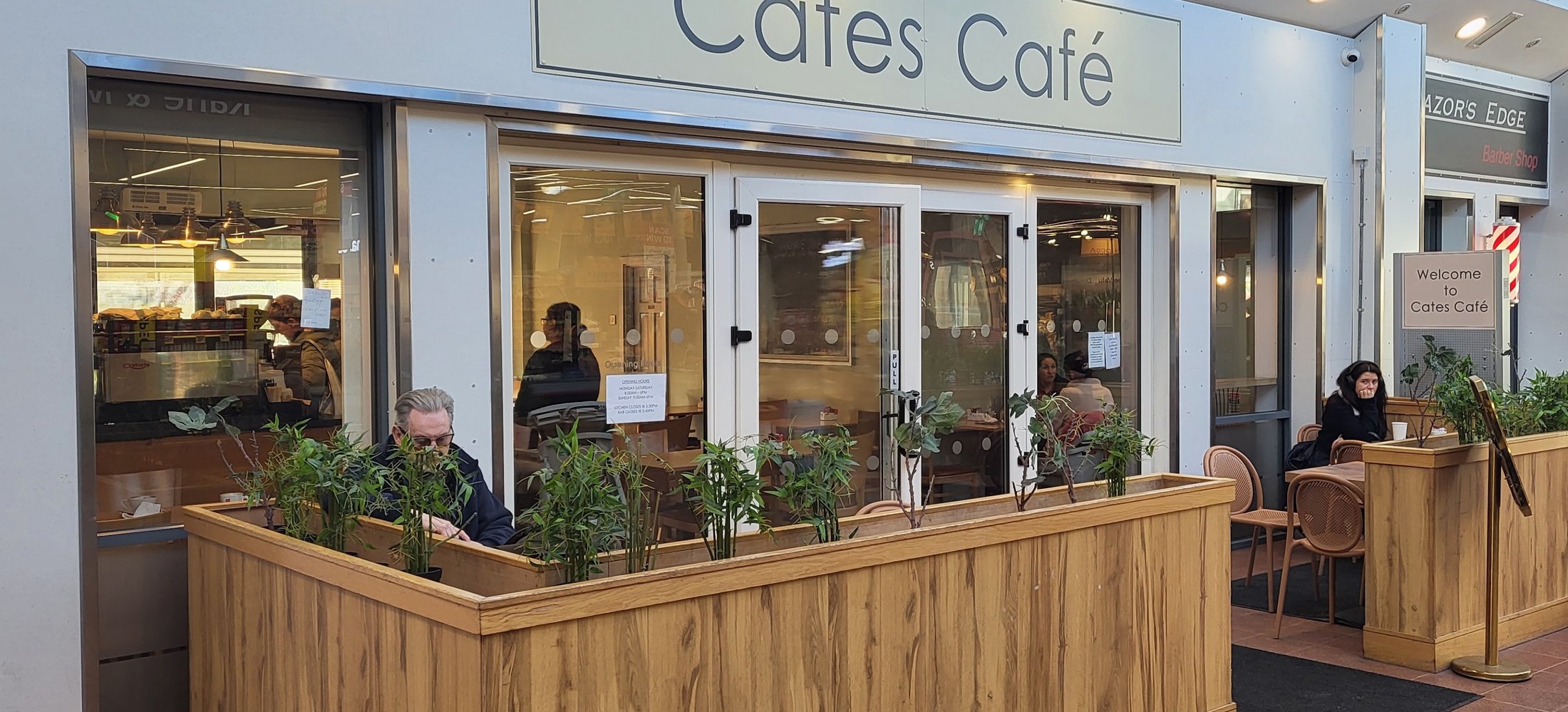 The front of Cates Café inside Donabate Town Centre: a pale sign reading Cates Café above a glazed frontage, with a long wooden planter of bamboo marking out the seating and a welcome board by the door.